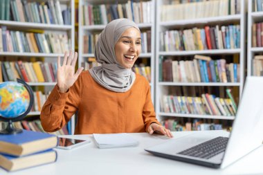 İyi stoklanmış bir kütüphanede kitaplarla çevrili dostane bir video görüşmesi sırasında dizüstü bilgisayarına tesettür dalgaları takan neşeli Müslüman kadın..