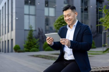 Asyalı gülümseyen genç bir adam, ofis merkezinin dışında oturuyor, kulaklık takıyor ve tablet üzerinde geziniyor..