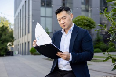 Ciddi bir Asyalı genç adam takım elbiseli bir ofis merkezinde oturuyor ve belgelere ve anlaşmaya bakıyor..