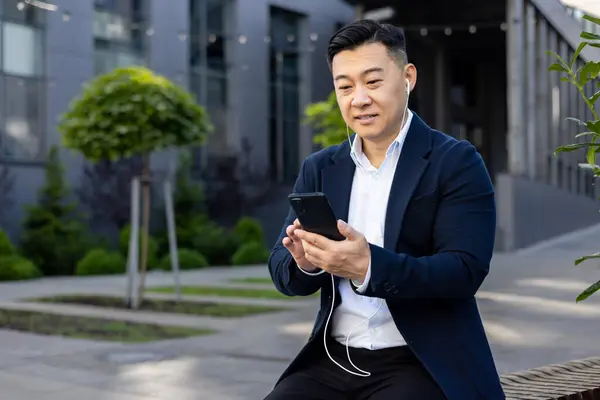 Asian young man in a business suit sitting outside on a bench near an office building wearing headphones and using a mobile phone.