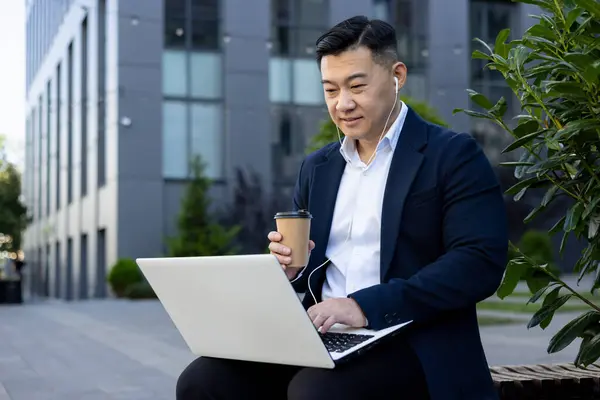 Asian young male businessman sitting on a bench near an office building wearing headphones, holding a cup of coffee in his hand and using a laptop.