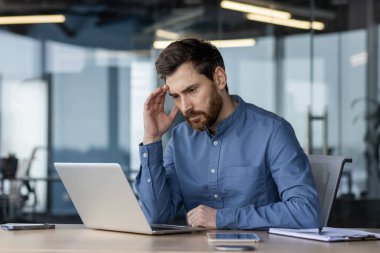 Ciddi ve düşünceli genç bir adam ofiste oturuyor, başını eline yaslıyor ve dizüstü bilgisayarın ekranına bakıyor, bir soruyu ve problemi çözüyor..