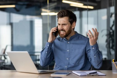 Sinirli ve sinirli genç iş adamı masanın başında oturuyor ve ellerini sallayarak telefonla konuşuyor..