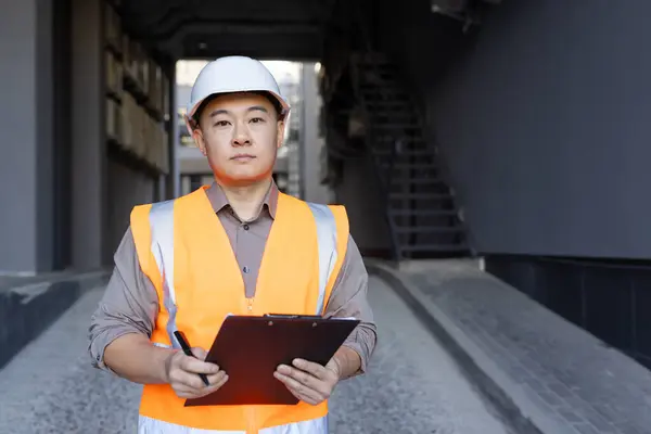 Portrait of serious and successful young Asian man in hard hat and vest standing outside building with folder and pen in hands, inspecting, developing project.