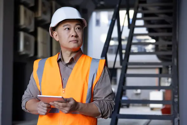 Photo of an Asian young male engineer, architect, foreman standing in a hard hat and vest outside the building, holding a tablet in his hands and looking confidently to the side.