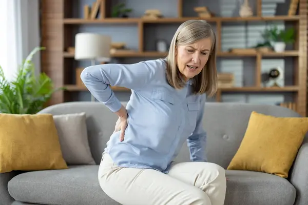 Unhealthy mature woman keeping hand on back while struggling with pain in spinal bones. Ill female experiencing problem with getting up from couch while squinting face from ache in flat interior.