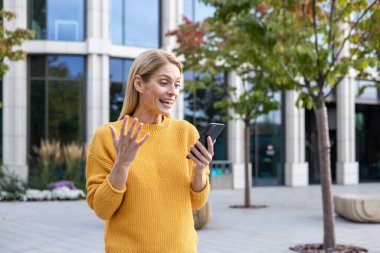 A joyful middle-aged woman in a bright yellow sweater uses her smartphone, visibly happy about the news she is receiving, standing in a modern urban park. clipart
