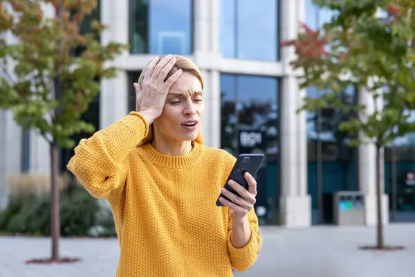 A middle-aged woman appears distressed and worried while she holds a smartphone on a sunny day with an urban background. Her expression conveys shock and confusion.