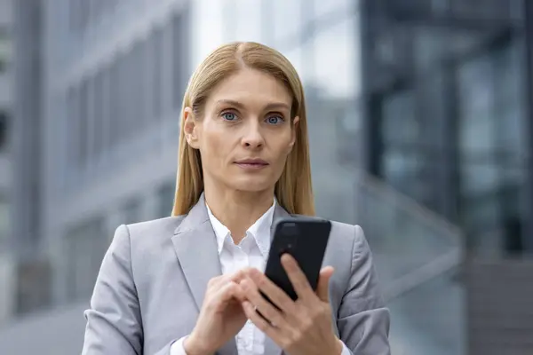 A confident businesswoman in a grey suit uses her smartphone against a modern urban background, conveying a sense of business communication and professionalism in a bustling city environment.