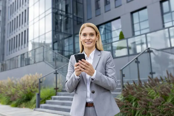 stock image A confident professional woman in a formal suit stands outside a contemporary office building, browsing her smartphone with a smile.