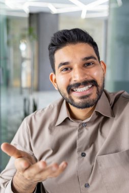 Sıradan tişörtlü, sakallı neşeli bir adam video görüşmesi yaparken el kol hareketi yapıyor. Kapalı alan çevresi modern ve iyi ışıklandırılmış. Görüntülü sohbet deneyimini ön plana çıkarıyor..