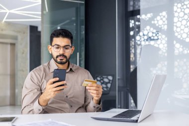 Modern bir ofiste genç bir işadamı akıllı telefonunu ve kredi kartını online alışveriş için kullanır. Ayarlar profesyonel ve teknoloji odaklı..