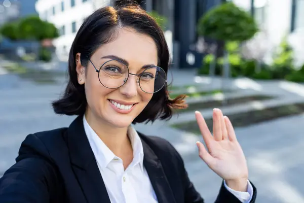 Smiling professional woman with glasses waving at the camera, making a friendly gesture during a video call outside her office building.