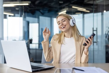 Spor ceketli neşeli genç bir kadın çalışma alanında müzik molası veriyor, kulaklık ve akıllı telefon kullanıyor, modern bir ofis ortamında keyif ve rahatlığı ifade ediyor..