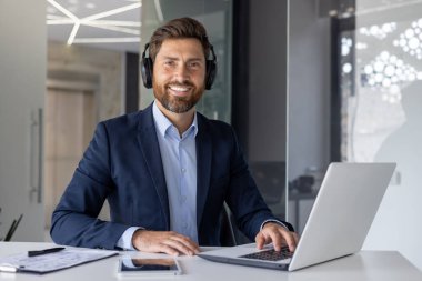 Takım elbiseli neşeli bir erkek profesyonel, şık bir ofis ortamında dizüstü bilgisayarda etkili bir şekilde çalışır. İletişim kurmak için kulaklık kullanır..