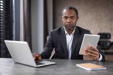 Modern ofis ortamında aynı anda bir laptop ve tablet kullanarak odaklanmış Afro-Amerikan iş adamı. Ciddi ve kendini işine adamış görünüyor..