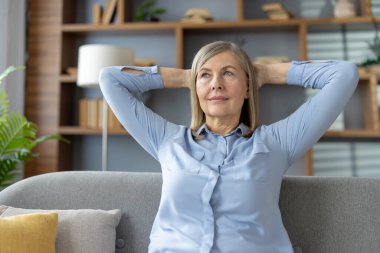 Huzurlu, kıdemli bir kadın evinde huzurlu bir anın tadını çıkarıyor. Elleri başının arkasında, kanepede oturuyor. Rahatlık ve memnuniyet hissini yansıtıyor..
