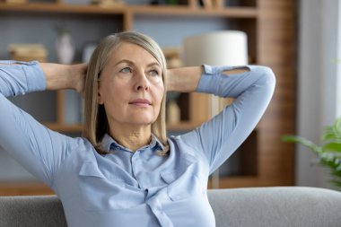 Mavi gömlekli olgun bir kadın ellerini başının arkasına koyup koltukta dinleniyor. Modern bir oturma odasında düşünceli ve sakin görünüyor..