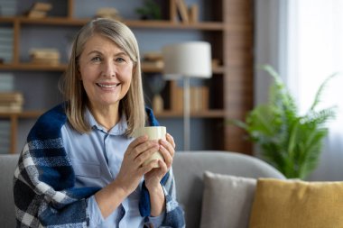 Ekoseli battaniyeye sarılı gülümseyen yaşlı bir kadın sıcak bir fincan kahve tutuyor ve şık oturma odasında huzurlu bir anın tadını çıkarıyor..