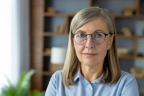 stock image A portrait of a confident senior woman wearing glasses and a light blue shirt, set in a modern office environment with a background of shelves.