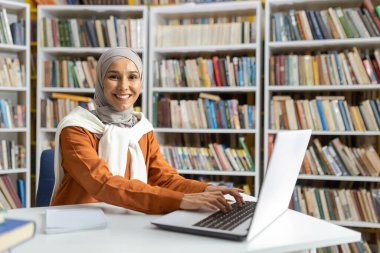 Tesettürlü neşeli Müslüman bir kadın kütüphanede bir masada oturuyor, dizüstü bilgisayarı üzerinde çalışıyor, dijital okuryazarlık ve eğitim gücünü sergiliyor..