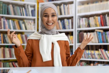 Tesettürlü neşeli Müslüman bir kadın, kütüphane ortamında kitaplarla çevrili bir video görüşmesi sırasında dostça bir jest yapıyor. Doğrudan kameraya bakıyor, sıcaklık ve pozitiflik yayıyor..