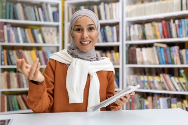 tesettürlü gülümseyen Müslüman kadın kütüphanede arkadaşça bir video görüşmesi yapıyor, doğrudan kameraya bakıyor, kitaplarla çevrili..