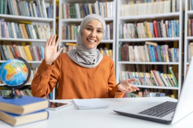 İnternette ders verirken tesettüre girmiş neşeli bir eğitimci. Etrafı kitaplarla ve dünyayla çevrili bir kütüphane ortamında misafirperver ve bilgili bir hava yayıyor..