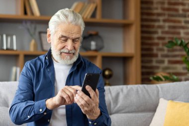 Beyaz sakallı yaşlı bir adam akıllı telefonunu kullanırken gülümsüyor, rahatça oturma odasında oturuyor, teknolojiyle rahatlık ve neşe gösteriyor..