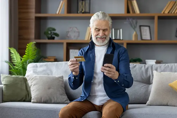 Cheerful senior man using smartphone and credit card for online shopping while sitting comfortably on sofa, in a well-decorated living room.
