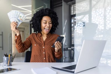 Hevesli Afrikalı Amerikalı iş kadını elinde nakit para tutuyordu ve modern ofis ortamında cep telefonuna heyecanla bakıyordu. Finansal başarı ve sevinç ifadesi.