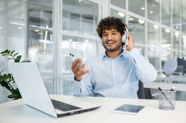 Genç gülümseyen adam, akıllı telefonuyla modern ofis ortamında iş yaparken müzikten zevk alıyor. İş hayatı dengesi ve teknoloji kavramı.