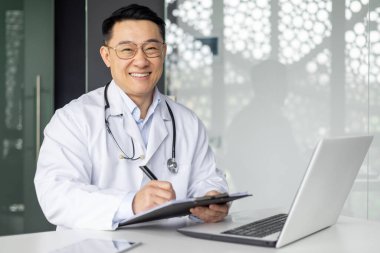 Neşeli Asyalı olgun erkek doktor ofisinde oturuyor, bir dizüstü bilgisayar üzerinde çalışıyor ve elinde bazı belgeler tutuyor, profesyonellik ve dostluk yayıyor..