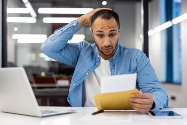 Şok olmuş genç bir Arap adam modern bir ofiste oturuyor ve bir zarfın içinde elini endişeyle kafasının arkasında tutarak bir mektup okuyor..