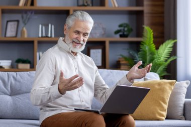 Gri saçlı, gülümseyen yaşlı adam dizüstü bilgisayarında video görüşmesi yapıyor. Evdeki kanepede oturuyor ve uzaktan konuşuyor..