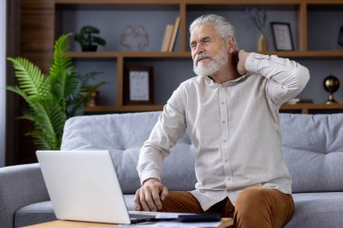 Yorgun, yaşlı, kır saçlı bir adam elinde dizüstü bilgisayarla koltukta oturur ve boynunu tutarak Asana 'yı yapar, dinlenir..