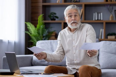 Evdeki kanepede oturan, dizüstü bilgisayarda çalışan, faturaları kontrol eden, makbuzları alan, kameraya bakıp ellerini kaldıran yaşlı, gri saçlı bir adamın portresi..