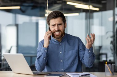Sinirli genç bir iş adamının portresi masada oturuyor ve duygusal olarak cep telefonuyla konuşuyor, ellerini sallıyor..