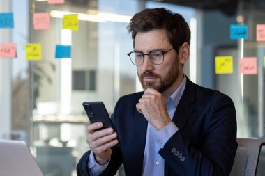 Ciddi düşünen bir iş adamı telefonda uygulama kullanıyor, takım elbiseli bir adam iş yerinde olgun bir patron, ofis içinde dizüstü bilgisayarı olan, yatırımcı çalışıyor..