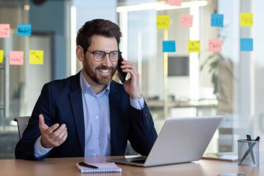 Takım elbiseli bir adam elinde dizüstü bilgisayarla masada otururken cep telefonuyla konuşuyor.