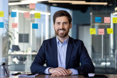 Sakallı ve kendine güveni olan profesyonel genç bir işadamı arka planda yapışkan notlarla dolu modern bir ofiste oturuyor..