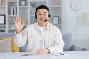 Kulaklık takan neşeli Asyalı bir adam kameraya gülümseyip el sallıyor, arka planda dekor olan evinde rahatça oturuyor..