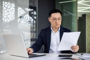 Takım elbiseli odaklı Asyalı bir işadamı çağdaş bir ofiste masasındaki belgeleri dikkatle inceliyor. Çevre şık ve aydınlık, profesyonel görünümünü geliştiriyor..