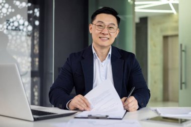 Modern bir ofis masasında çalışırken gülümseyen kendine güvenen Asyalı işadamı elinde dizüstü bilgisayar ve kalemle belgeleri inceliyor..