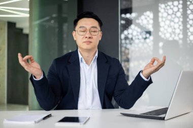 Modern bir ofiste masasında meditasyon yapan profesyonel bir iş adamı. Yoğun bir çalışma ortamında stresi yönetmek ve odaklanmayı artırmak için farkındalık uyguluyor..