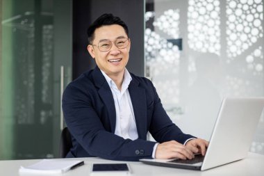 Neşeli Asyalı iş adamı, çağdaş ofis ortamında bilgisayarını kullanarak başarıyı ve profesyonelliği resmediyor.