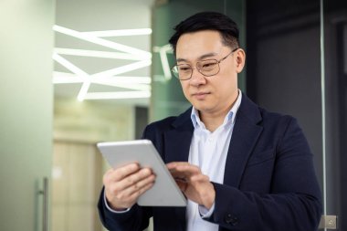 Kendine güvenen Asyalı işadamı modern iş ve teknoloji kullanımını yansıtan modern neon ışıkları altında çağdaş bir ofis ortamında dijital tablet kullanıyor..