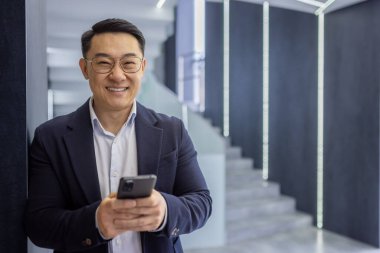 Takım elbiseli kendine güvenen Asyalı işadamı şık iç mekanları olan şık, çağdaş ofislerde akıllı telefonunu kullanırken sıcak bir şekilde gülümsüyor..