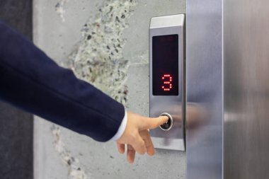 İyi giyimli bir işadamı asansör düğmesine basmak için çağdaş bir ofis binasında üçüncü kata sinyal gönderiyor. Görüntü, iş dünyasındaki teknoloji ve erişilebilirliği yakalıyor..