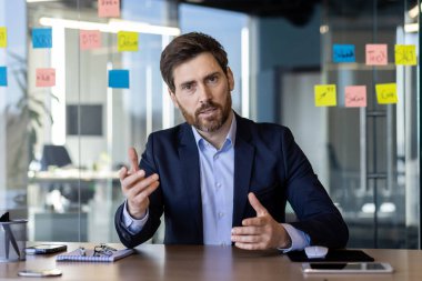 Modern bir ofis ortamında video aracılığıyla iletişim kuran odaklanmış işadamı, eliyle işaret ederken önemli konuları tartışıyor..